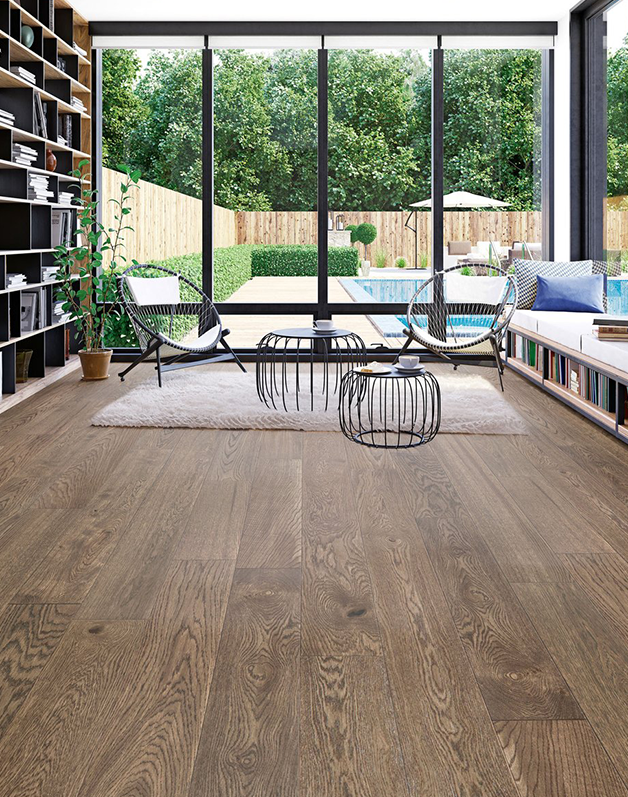 A modern living room with large floor-to-ceiling windows overlooking a backyard pool and patio area. The room features wide-plank hardwood floors with a natural, matte finish, light brown in tone. The space is furnished with minimalist black wire-frame chairs, a long low-shelf bookcase, and a built-in wall bookshelf with books and decorative items. The room is bright, with natural light streaming in through the windows, and the view outside includes a well-maintained lawn, wooden fencing, and patio furniture