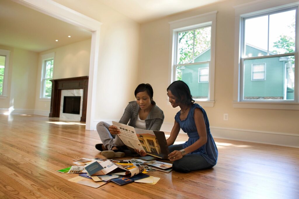 Two women sitting on a hardwood floor, reviewing brochures and design ideas in a bright, spacious room